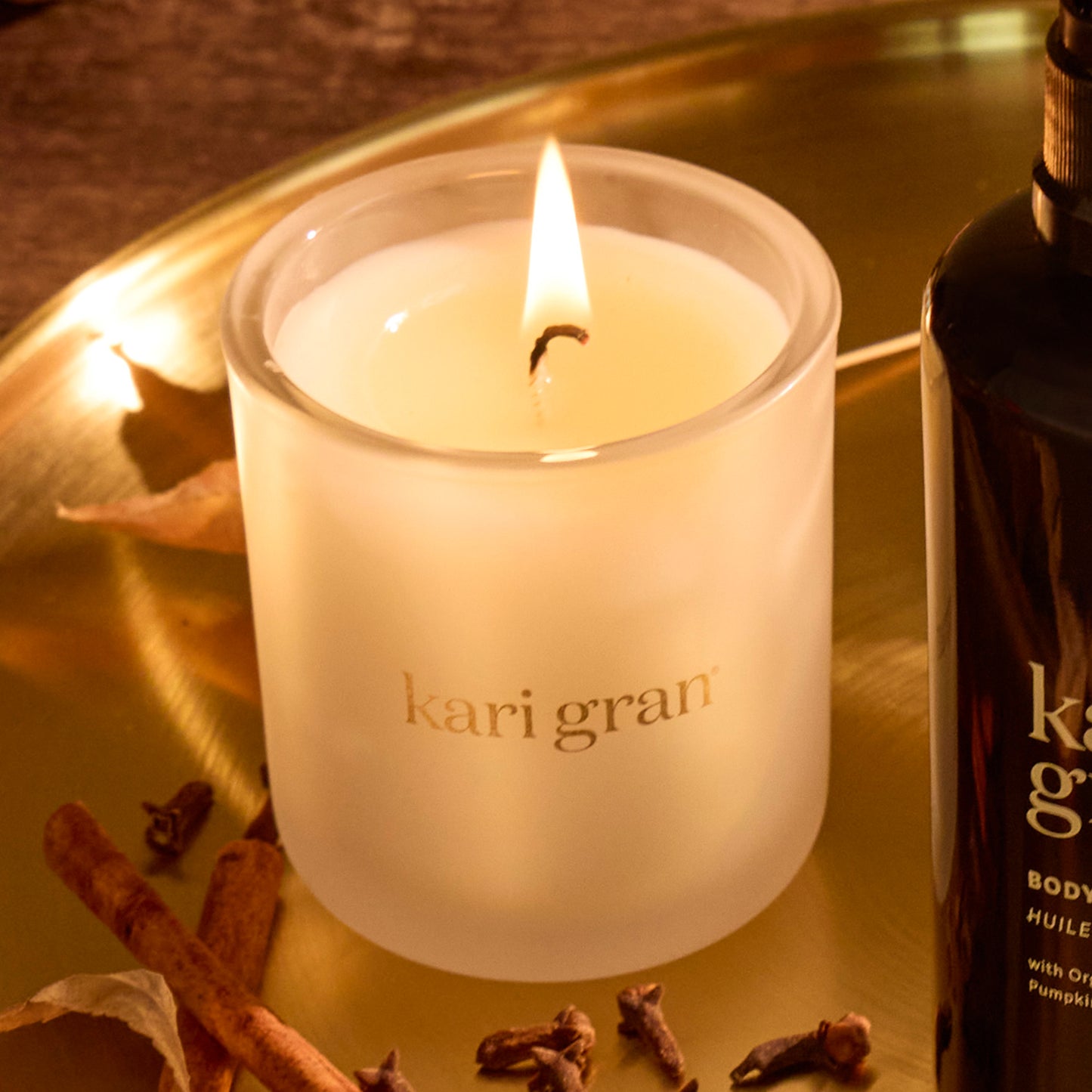 Kari gran candle and bottle on a reflective surface with cinnamon sticks and cloves.