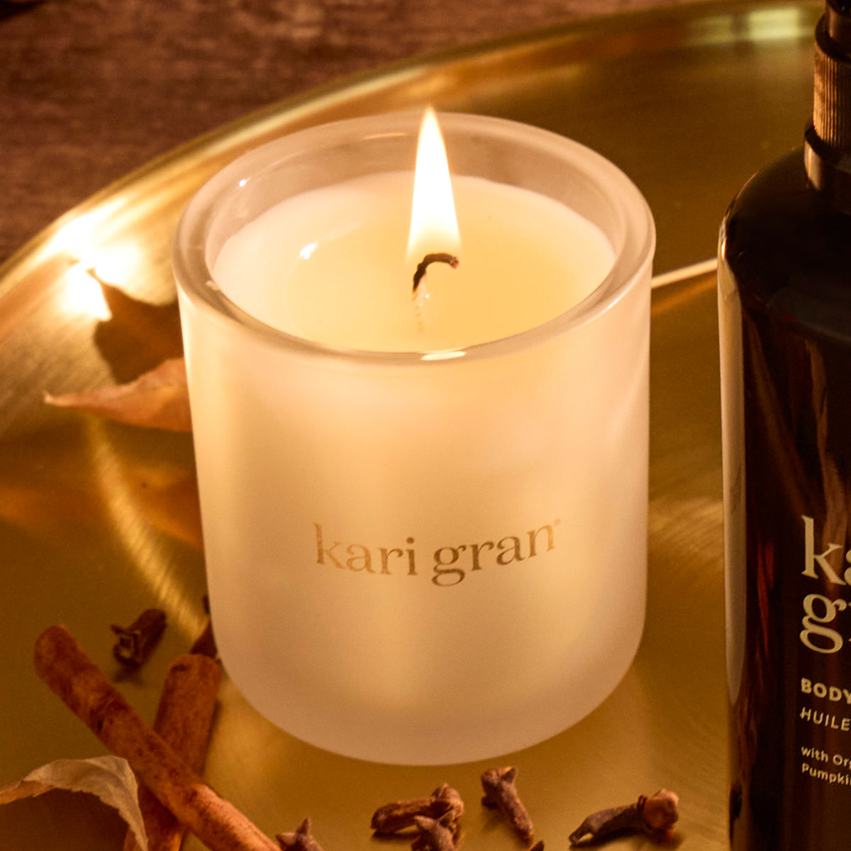 Kari gran candle and bottle on a reflective surface with cinnamon sticks and cloves.